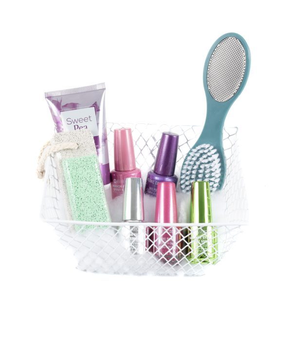 The Manicure/Pedicure Basket includes a white wire basket that holds a tube of Sweet Pea lotion, a green pumice stone with an attached rope, an assortment of colorful nail polishes, and a dual-sided foot scrubbing brush with a teal handle. Perfect for both pedicures and manicures, the basket is lined with white tissue paper.