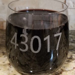 A clear glass filled with dark liquid, possibly red wine, sits on a granite countertop. The number 43017, a zip code, is etched prominently on the side of the glass. In the background, a tiled wall and a pair of rocks/juice glasses from the "Pair of Rocks/Juice Glasses - Choose any Zip Code" collection are nearby.