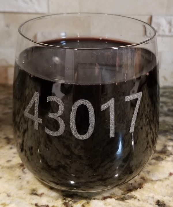 A clear glass filled with dark liquid, possibly red wine, sits on a granite countertop. The number 43017, a zip code, is etched prominently on the side of the glass. In the background, a tiled wall and a pair of rocks/juice glasses from the "Pair of Rocks/Juice Glasses - Choose any Zip Code" collection are nearby.