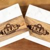 Two square coasters placed on a wooden surface. Each coaster, named "Pair Square Coasters - Your Custom Text/Logo Here," features a wooden center with the Texas Roadhouse logo etched into it, flanked by white marble on the top and bottom. These custom logo coasters add a personalized touch to any setting.