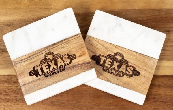 Two square coasters placed on a wooden surface. Each coaster, named "Pair Square Coasters - Your Custom Text/Logo Here," features a wooden center with the Texas Roadhouse logo etched into it, flanked by white marble on the top and bottom. These custom logo coasters add a personalized touch to any setting.