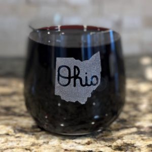 A stemless wine glass from the "Pair of Rocks/Juice Glasses - State of Ohio with Script Ohio" set, filled with a dark beverage, sits on a granite countertop. The glass showcases a graphic of the state of Ohio with "Ohio" inscribed inside the state outline, reminiscent of Script Ohio. The background is softly blurred.