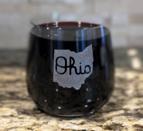 A stemless wine glass from the "Pair of Rocks/Juice Glasses - State of Ohio with Script Ohio" set, filled with a dark beverage, sits on a granite countertop. The glass showcases a graphic of the state of Ohio with "Ohio" inscribed inside the state outline, reminiscent of Script Ohio. The background is softly blurred.