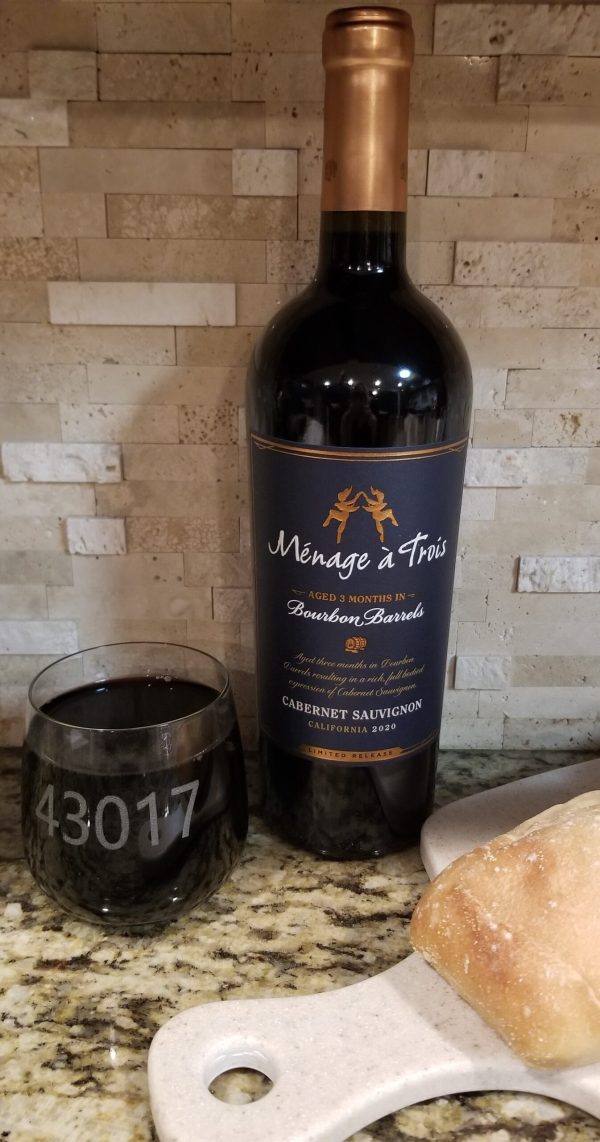 A bottle of Ménage à Trois Cabernet Sauvignon wine stands on a kitchen countertop beside a piece of bread on a cutting board and a pair of rocks/juice glasses, custom-engraved with your preferred text or logo. The bottle label mentions it is aged in bourbon barrels. The background features a tiled backsplash.