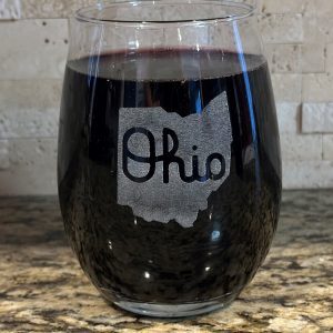 A stemless wine glass from the "Pair of Wine Glasses - State of Ohio with Script Ohio" set, filled with dark red wine and showcasing an outline of the state of Ohio with the word "Ohio" elegantly scripted inside it, is placed on a granite countertop with a stone-tiled backsplash in the background.