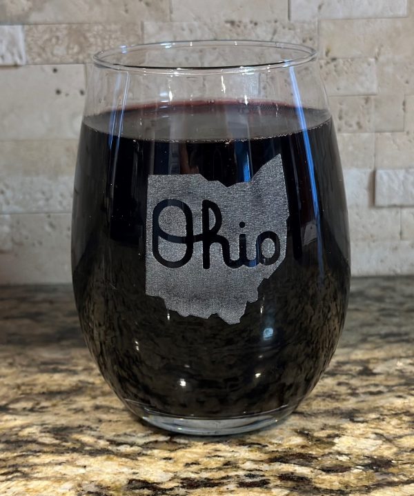 A stemless wine glass from the "Pair of Wine Glasses - State of Ohio with Script Ohio" set, filled with dark red wine and showcasing an outline of the state of Ohio with the word "Ohio" elegantly scripted inside it, is placed on a granite countertop with a stone-tiled backsplash in the background.