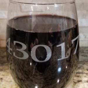 A pair of **Pair of Wine Glasses - Choose any Zip Code**, each etched with the zip code "43017" in large, clear fonts, are placed on a granite countertop. The background showcases a light stone backsplash with subtle patterns.