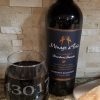 A bottle of Ménage à Trois Cabernet Sauvignon aged 3 months in bourbon barrels stands next to one of our wine glasses engraved with the zip code "43017." Below them lies a cutting board with a piece of crusty bread, all set on a granite countertop with a stone-tiled backsplash.