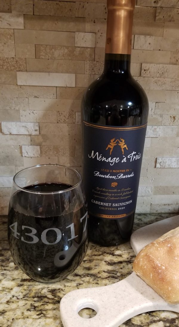 A bottle of Ménage à Trois Cabernet Sauvignon aged 3 months in bourbon barrels stands next to one of our wine glasses engraved with the zip code "43017." Below them lies a cutting board with a piece of crusty bread, all set on a granite countertop with a stone-tiled backsplash.