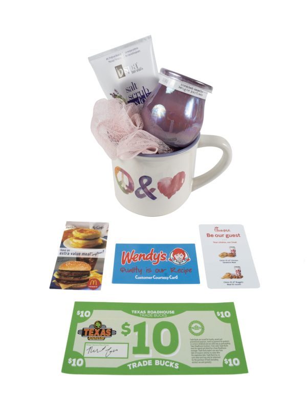 The Peace & Love Mug Gift Basket with Free Food Certificate features a colorful, stylized "Q" and heart symbol on the mug. Inside, you'll find a pink loofah, hand cream, and a purple jar. Accompanying the mug are gift cards for McDonald's, Wendy's, Chick-fil-A, and a $10 Texas Roadhouse Free Food Certificate.