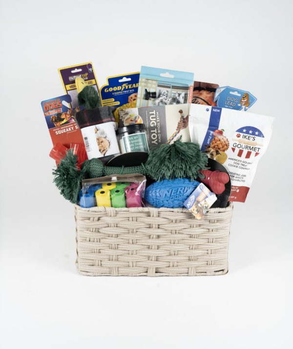 The Adult Gambling Night Basket is a beige woven basket filled with various dog toys and treats, perfect for an adult night in with your furry friend. Items include dog snacks, chew toys, a rope toy, a rubber ball, and packages of different dog-friendly foods and treats. The basket is neatly arranged with colorful packaging and toys visible.