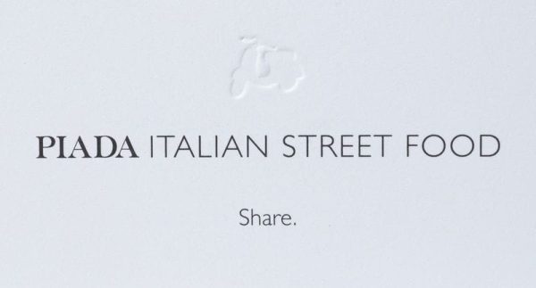 A white sign features the embossed outline of a scooter at the top. Below, black capital letters read "PIADA ITALIAN STREET FOOD," with the word "Share." centered in smaller black font underneath. Enjoy our protein-packed dishes or grab Piada BOGO Cards (includes protein) for your next meal!