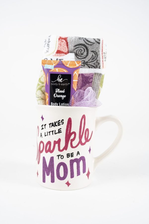 The Sparkle Mom Mug Gift Basket with Free Food Certificate includes a white ceramic mug adorned with the phrase "It takes a little sparkle to be a Mom" in pink and purple letters. The mug comes filled with a rolled colorful towel, a tube of blood orange body lotion, and a purple loofah mesh.