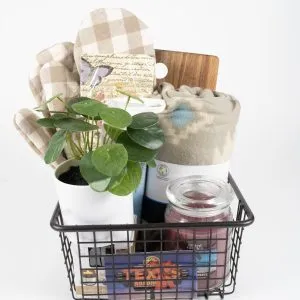 A Texas Road House Kitchen & Home Basket, containing $75 in Texas Roadhouse Certificates, features a metal basket with a potted plant, plaid oven mitts, a small wooden board, a rolled blanket, a large red candle, and a white candle. The items are all neatly arranged against a white background.