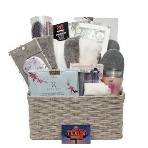 The Deluxe Spa Basket - Includes $75 in Texas Roadhouse Certificates, is filled with various spa and comfort items such as a sleep mask, cozy socks, a hairbrush, skincare products, a candle, and an eye gel mask. The items are assorted in neutral and calming colors.