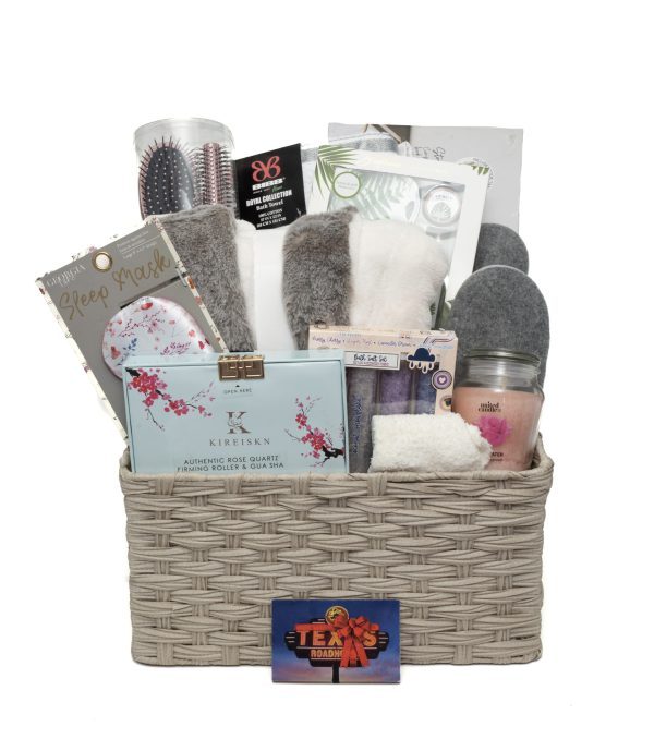 The Deluxe Spa Basket - Includes $75 in Texas Roadhouse Certificates, is filled with various spa and comfort items such as a sleep mask, cozy socks, a hairbrush, skincare products, a candle, and an eye gel mask. The items are assorted in neutral and calming colors.