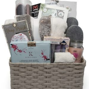 The Deluxe Spa Basket - Our Largest Spa Basket, features a beige wicker basket filled with an array of spa and comfort items including a sleep mask, candle, rose quartz roller, fluffy socks, slippers, bath brush, hairbrush, tea, body wash, and a bathrobe. These items are neatly arranged to create the perfect indulgent gift.