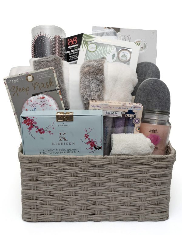 The Deluxe Spa Basket - Our Largest Spa Basket, features a beige wicker basket filled with an array of spa and comfort items including a sleep mask, candle, rose quartz roller, fluffy socks, slippers, bath brush, hairbrush, tea, body wash, and a bathrobe. These items are neatly arranged to create the perfect indulgent gift.