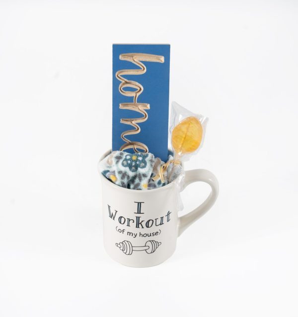 The "I Work Out of the House Mug Gift Basket with Free Food Certificate" includes a white ceramic mug featuring the humorous text "I Workout (of my house)" accompanied by a dumbbell icon. The basket also contains a fabric item adorned with a floral pattern, a cheerful yellow lollipop, and a blue card inscribed with the word "home" in gold.