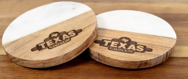 Two round coasters with white marble tops are pictured on a wooden surface. Both personalized coasters feature the Texas Roadhouse logo, part of the "Pair Round Coasters - Your Custom Text/Logo Here" product collection, engraved in the wood section.