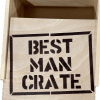 A light-toned wooden box with its lid slightly open. The lid, positioned to the side, has "BEST MAN CRATE" stenciled in bold, dark letters. Inside, while currently empty, it's designed to hold best man items such as a blue distressed hat.