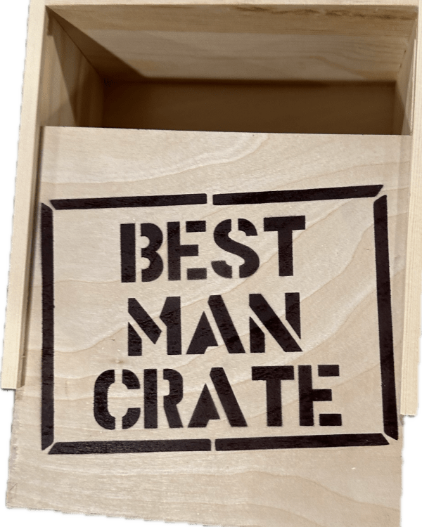 A light-toned wooden box with its lid slightly open. The lid, positioned to the side, has "BEST MAN CRATE" stenciled in bold, dark letters. Inside, while currently empty, it's designed to hold best man items such as a blue distressed hat.