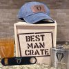 A display of Best Man themed items includes a crate labeled "Best Man Crate" containing four items: a blue distressed hat with a "Best Man" logo, a beer glass with "Best Man" on it, a black bottle opener featuring the same logo, and a metallic flask also labeled "Best Man.