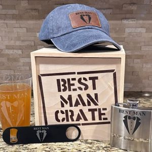 A display of Best Man themed items includes a crate labeled "Best Man Crate" containing four items: a blue distressed hat with a "Best Man" logo, a beer glass with "Best Man" on it, a black bottle opener featuring the same logo, and a metallic flask also labeled "Best Man.