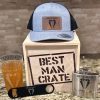 A "Best Man Crate with 4 Best Man Items including Gray Snapback Hat" is displayed on a countertop. The set includes a gray snapback hat with a tuxedo emblem, a wooden crate labeled "Best Man Crate," a pint glass with "Best Man" text, a bottle opener, and a flask, all featuring the same tuxedo logo.