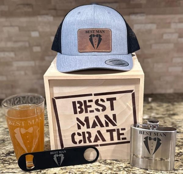 A "Best Man Crate with 4 Best Man Items including Gray Snapback Hat" is displayed on a countertop. The set includes a gray snapback hat with a tuxedo emblem, a wooden crate labeled "Best Man Crate," a pint glass with "Best Man" text, a bottle opener, and a flask, all featuring the same tuxedo logo.