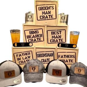 A display of wooden crates labeled for various wedding roles, such as "Groom's Man," "Ring Bearer," and "Father in Law." One crate is the Best Man Crate with 4 Best Man Items, including a blue distressed hat. In front of the crates are caps and drinkware with matching labels. Additionally, there is a bottle opener, flasks, and a pint glass included.