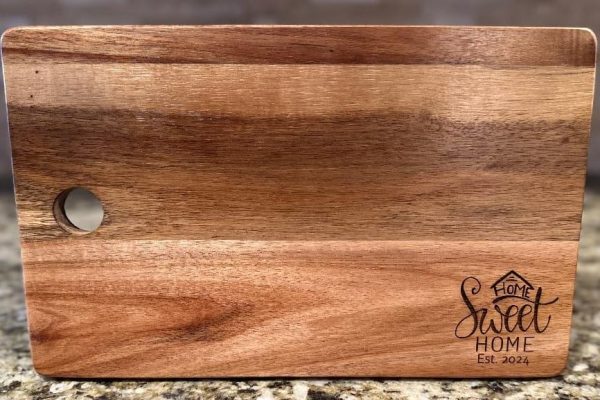 The Cutting Board - Home Sweet Home 2024, a rectangular wooden cutting board with a smooth, polished surface, is displayed on a granite countertop. It features a round hole in the top left corner and an engraving in the bottom right that reads "Home Sweet Home Est. 2024," complete with a small house icon above the text.