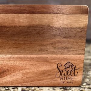 Introducing the Cutting Board - Home Sweet Home 2024: a wooden cutting board with a smooth finish and a sophisticated two-tone color palette. The bottom-right corner showcases an elegantly engraved inscription, "Home Sweet Home Est. 2024," accompanied by a small house illustration. This charming cutting board rests beautifully on any speckled countertop, adding a touch of warmth and style to your kitchen décor.