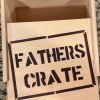 A "Fathers Crate - Wooden Gift Box" with an open lid sits on a granite countertop. The inside of the lid has the words "FATHERS CRATE" stenciled in bold black letters within a rectangular border. The box appears to be empty.