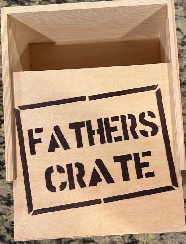 A "Fathers Crate - Wooden Gift Box" with an open lid sits on a granite countertop. The inside of the lid has the words "FATHERS CRATE" stenciled in bold black letters within a rectangular border. The box appears to be empty.