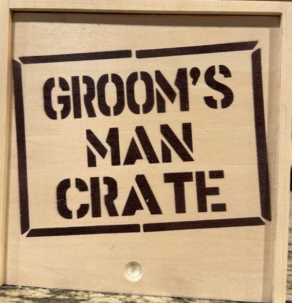 A Groom Man Crate - Wooden Gift Box with the text "GROOM'S MAN CRATE" stenciled in bold, black letters on the front. The crate features a natural wood finish and is displayed on a speckled surface.