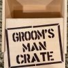 The Groom Man Crate - Wooden Gift Box, with its lid partially open to reveal an empty interior, displays the text "GROOM'S MAN CRATE" in bold, black stencil lettering on the lid. The box is situated on a granite countertop.