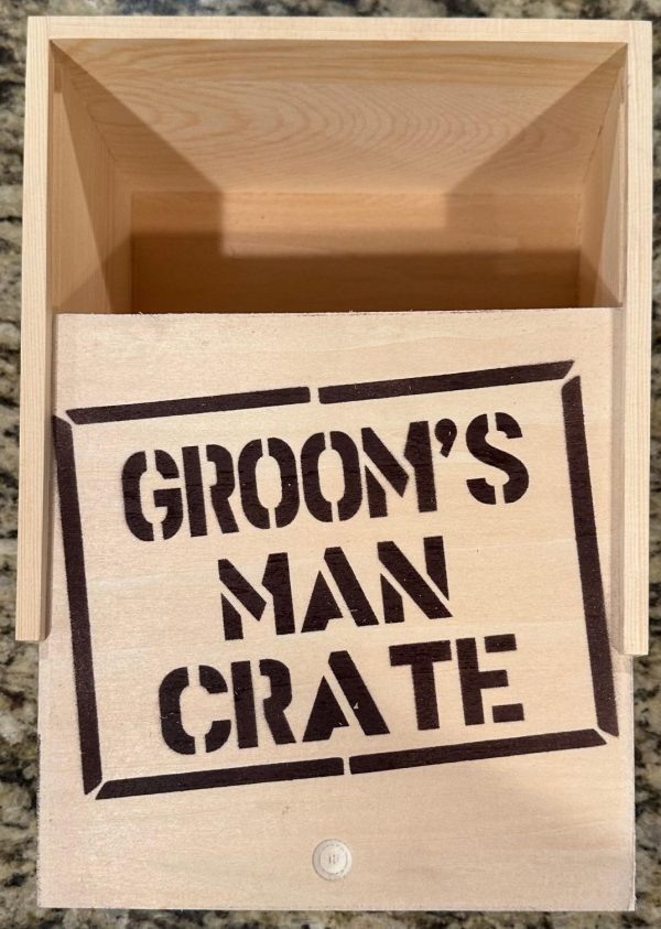 The Groom Man Crate - Wooden Gift Box, with its lid partially open to reveal an empty interior, displays the text "GROOM'S MAN CRATE" in bold, black stencil lettering on the lid. The box is situated on a granite countertop.