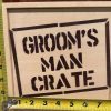 A product labeled "Groom Man Crate - Wooden Gift Box" is showcased with two yellow measuring tapes positioned across it. One tape measures the height, showing approximately 9 inches, and the other measures the width, showing about 12 inches.