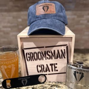 A wooden crate labeled "GROOMSMAN CRATE" sits on a countertop. Surrounding it are various groomsman items from the set: a blue distressed hat with a "Groomsman" leather patch, a pint glass featuring "Groomsman" and a tuxedo icon, a black bottle opener, and a metal flask with matching branding.