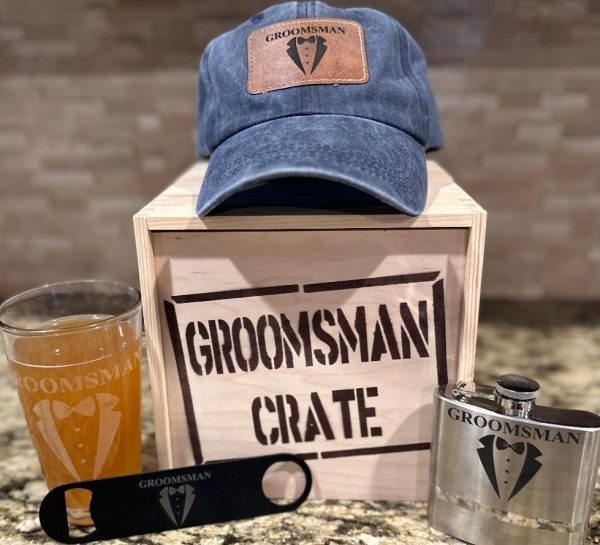 A wooden crate labeled "GROOMSMAN CRATE" sits on a countertop. Surrounding it are various groomsman items from the set: a blue distressed hat with a "Groomsman" leather patch, a pint glass featuring "Groomsman" and a tuxedo icon, a black bottle opener, and a metal flask with matching branding.