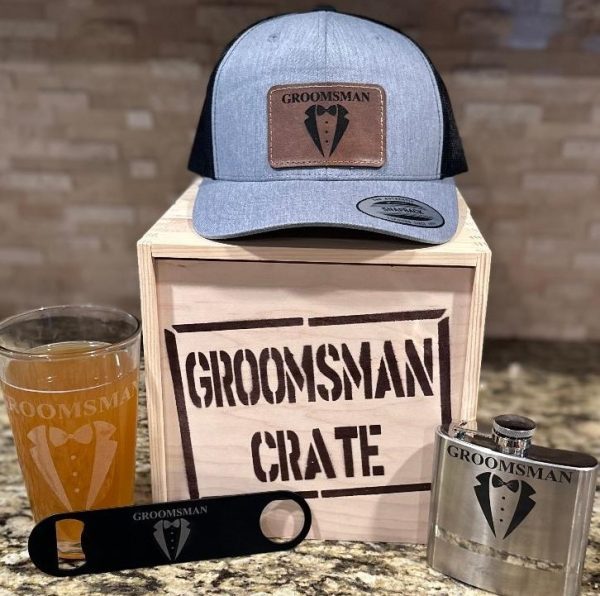 The image showcases the Groomsman Crate with 4 Groomsman Items including a Gray Snapback Hat. Inside the wooden crate labeled "GROOMSMAN CRATE," you'll find a gray snapback hat featuring a "GROOMSMAN" logo, a glass filled with beer, a flask, and a bottle opener. Each piece in this collection sports matching tuxedo-themed designs, making it an ideal gift for groomsmen.