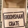 A Groomsman Crate - Wooden Gift Box with an open lid labeled "Groomsman Crate" in bold, black letters sits prominently on a granite countertop, appearing empty inside.