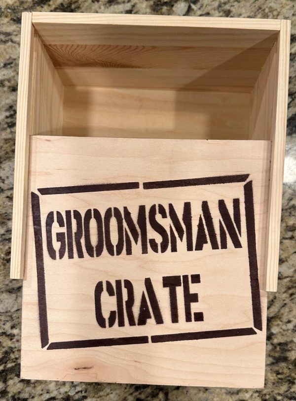 A Groomsman Crate - Wooden Gift Box with an open lid labeled "Groomsman Crate" in bold, black letters sits prominently on a granite countertop, appearing empty inside.