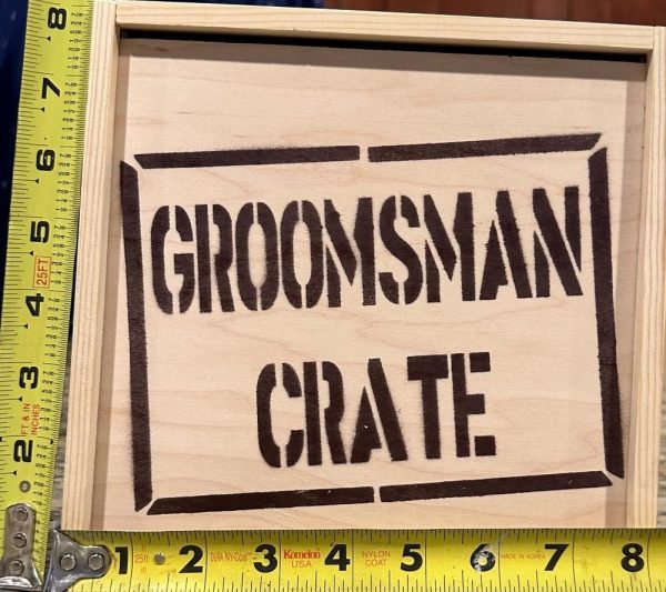 The Groomsman Crate - Wooden Gift Box features "GROOMSMAN CRATE" stenciled in black letters on its lid. Measured with a yellow tape measure, it shows approximately 10 inches in width and 7.5 inches in height, making it the perfect size for any groomsman's essentials.