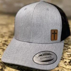 The Classic Snapback Hat with Leatherette Cross patch, featuring a gray and black design, displays a front leatherette cross patch and includes a brim sticker reading "The Authentic Yupoong Snapback." This stylish hat is showcased on a granite surface.