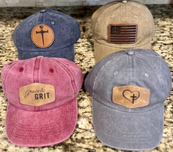 Four Faith Based Hats are displayed on a granite countertop, arranged two in front and two in back. The front left hat is red with a "grace & GRIT" label, the front right hat is grey featuring a heart and cross patch. The back left hat is blue with a cross emblem, and the back right hat is beige adorned with an American flag patch.