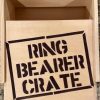 A Ring Bearer Crate - Wooden Gift Box, prominently labeled with bold, black stencil-style letters on the lid, sits open to reveal its empty interior. The crate rests on a surface with a granite-like texture visible in the background.