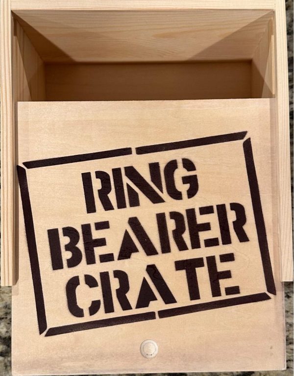 A Ring Bearer Crate - Wooden Gift Box, prominently labeled with bold, black stencil-style letters on the lid, sits open to reveal its empty interior. The crate rests on a surface with a granite-like texture visible in the background.