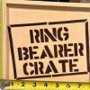 A "Ring Bearer Crate - Wooden Gift Box" with the name stenciled on the front is being measured with two yellow tape measures, one for height and the other for width.